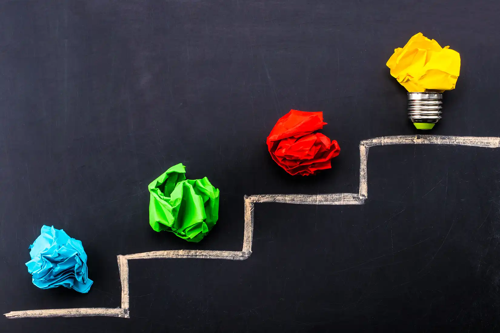 Evolving idea concept with colorful crumpled paper and light bulb on steps drawn on blackboard.